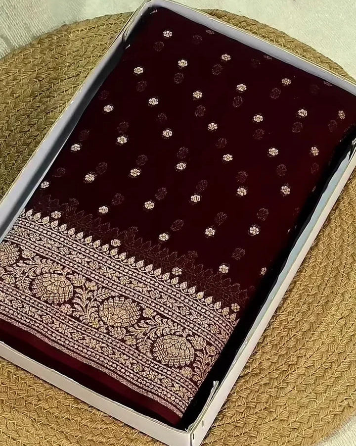 A dark maroon velvet fabric, likely a saree, displayed inside its clear plastic packaging. The fabric features small metallic gold floral motifs and a detailed gold zari border.