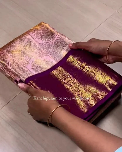 A person's hands holding and unfolding a traditional Indian saree, showing its rich purple body and intricate gold zari metallic border patterns, set against a light wood floor background.