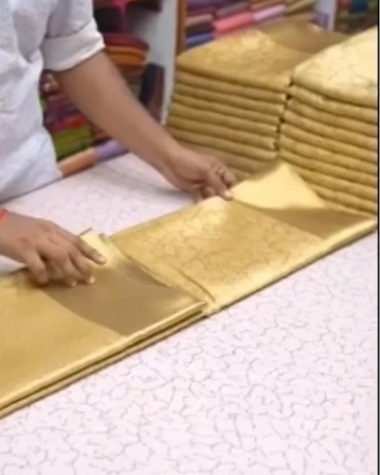 A person's hands and arms are visible as they unfold a gold-colored brocade fabric from a stack on a white patterned table. More stacks of folded gold fabric are visible in the background.