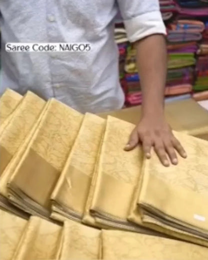 A person in a white shirt holds a stack of folded beige silk sarees with a subtle floral pattern, with a shop background showing other colorful folded fabrics.