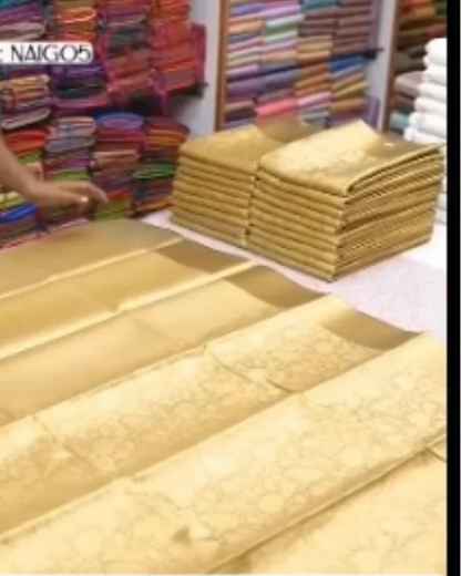 A gold-colored silk saree with a subtle floral pattern is draped on a table in a fabric store, with stacks of folded colorful textiles in the background.