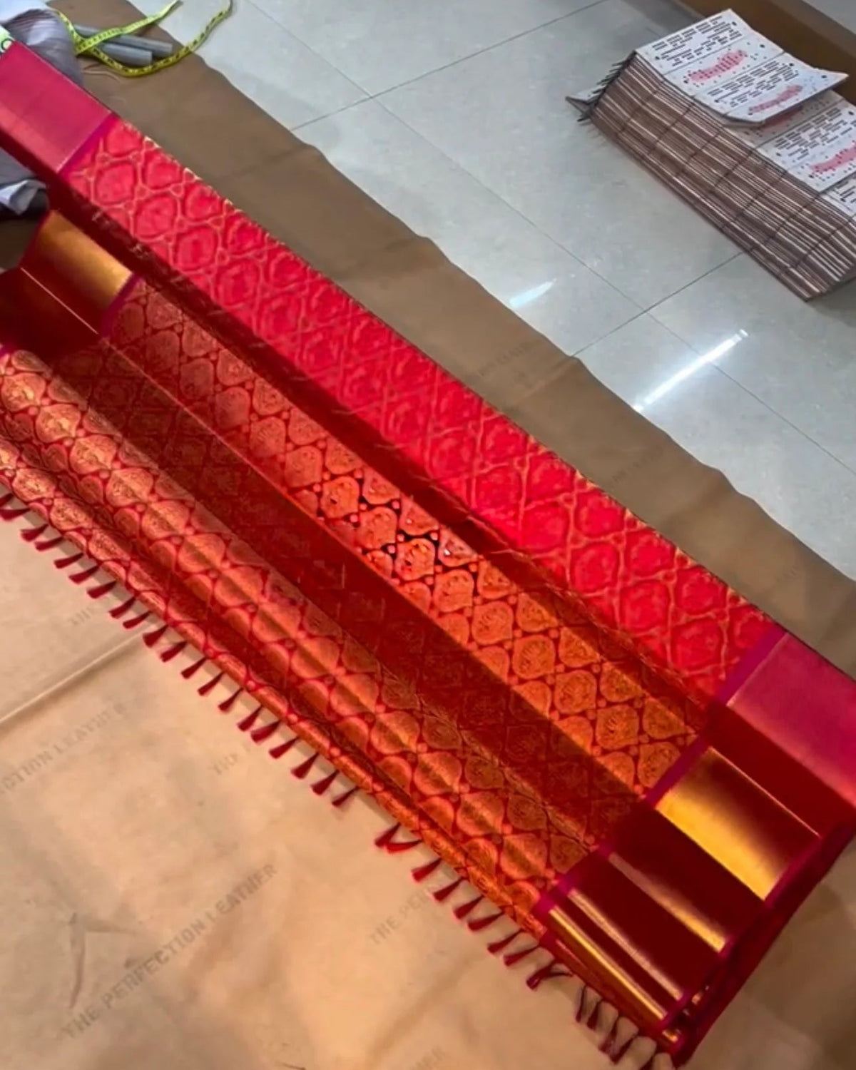A red and gold patterned leather belt with fringe detailing, laid diagonally across a light-colored floor.