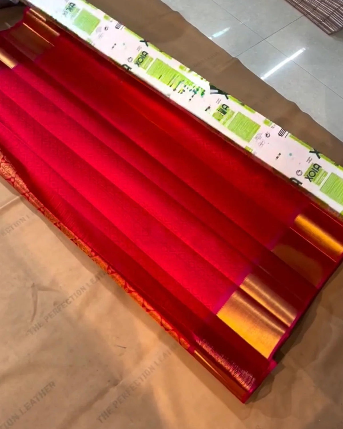 a roll of shiny red vinyl wrap or contact paper with a metallic gold edge, shown partially unrolled on a brown cardboard sheet on a tiled floor.