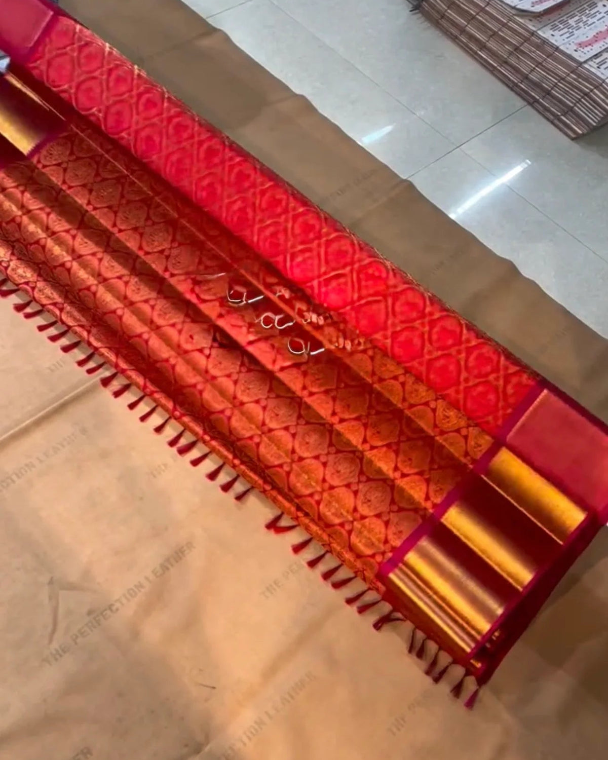 a close-up of a red and gold patterned fabric, likely a saree or scarf, laid out on a light-colored floor with a stack of similar fabrics in the background.